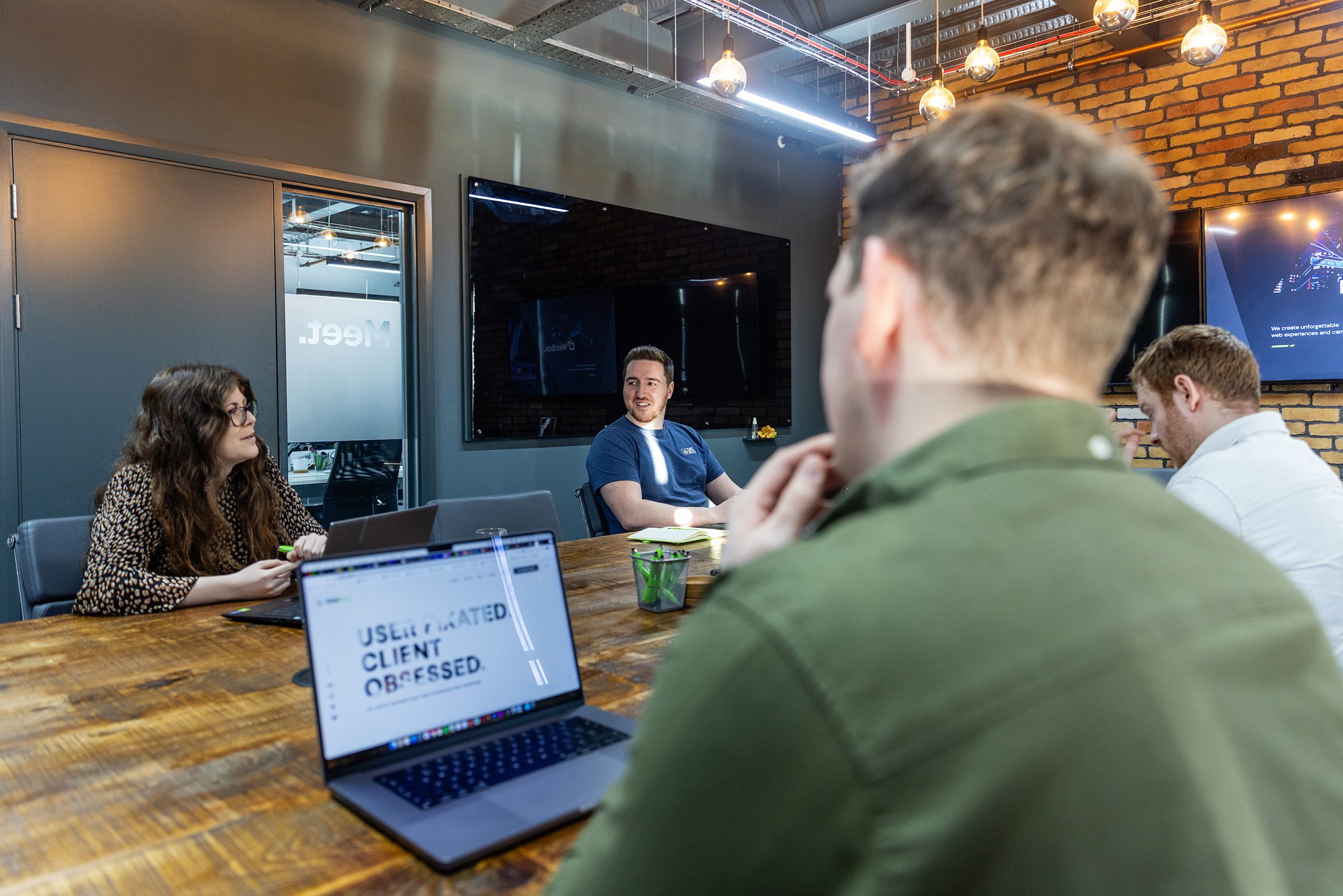 A group of 4 colleagues sit around a wooden table in a modern office meeting room, discussing work. Laptops are open on the table, and one screen displays text. The room features exposed brick walls, hanging lights, and wall-mounted screens.