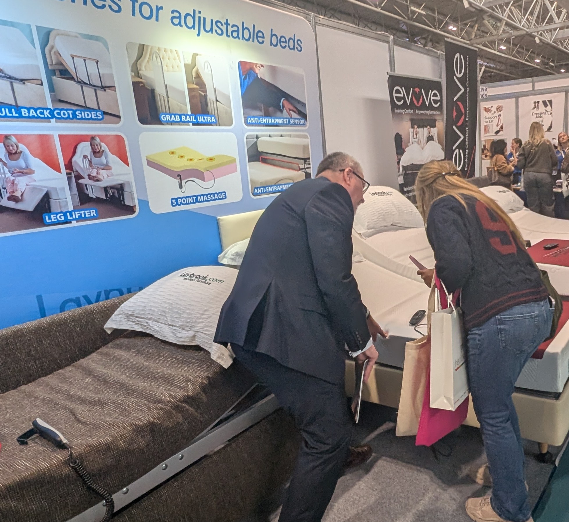 Two attendees at an exhibition stand look at and test adjustable beds, while a display board behind them highlights features such as grab rails, leg lifters, and massage functions.